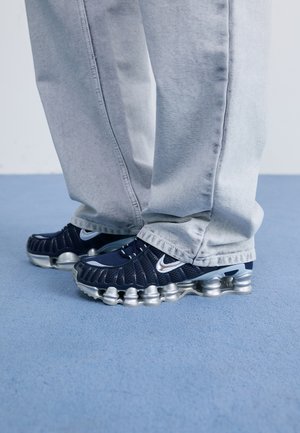 Marineblauwe Nike sneakers met een metallic zilveren zool, met een bovenwerk van mesh en opvallende ventilatie-details. Gekozen in combinatie met lichtgrijze broek.