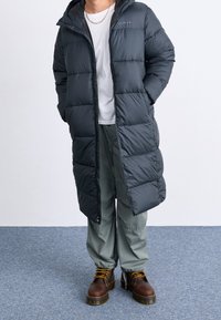 Mörkgrå puffjacka med huva, med quiltad vaddering, dragkedjestängning och sidofickor, som matchas med ljusgröna byxor och bruna stövlar.