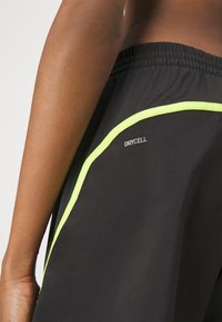 Pantalones cortos deportivos negros con textura de malla, que presentan un acento amarillo neón a lo largo del costado y un logo "DRYCELL" cerca de la cintura.