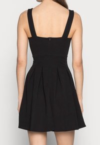 Svart ärmlös klänning med en åtsittande bodice, plisserad kjol och dragkedja i ryggen. Materialet har en slät textur och banden är breda.