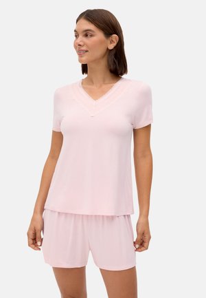 Mujer con blusa rosa claro de manga corta y escote en V con ribete de encaje y pantalones cortos sueltos a juego, de pie contra un fondo liso.