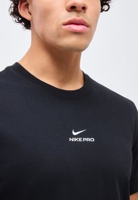 Νέος/Νεαρό άτομο που φοράει μαύρο μπλουζάκι Nike Pro με λευκό λογότυπο στο στήθος, αποκομμένο από τα χείλη μέχρι το πάνω μέρος του κορμού σε μια απλή φόντο.