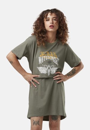 Femme aux cheveux bouclés portant une robe T-shirt graphique de moto Von Dutch vert olive avec des tatouages sur les bras et les jambes, mains sur les hanches.
