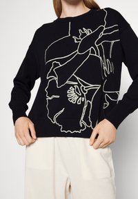 Czarny dzianinowy sweter z dużym abstrakcyjnym białym rysunkiem kwiatowym, noszony z jasnobeżowymi spodniami, ręce trzymające brzeg swetra.