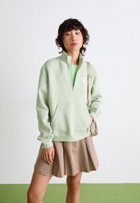 Sweatshirt zippé vert clair avec une poche avant et des poignets côtelés, associé à une jupe plissée beige et un petit sac. Texture en coton lisse.