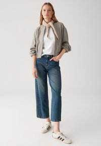 Giacca bomber corta grigio chiaro con maniche arricciate, abbinata a jeans dritti a vita alta blu medio e sneakers bianche con strisce nere.