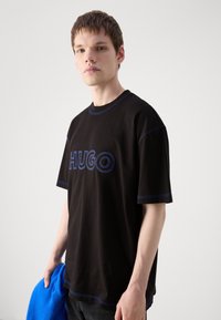 Melnas T-kreklas ar zilu kontrastējošu šuvumu un "HUGO" logo priekšpusē. Modelis tur zilu vilnas jaku. Vienkāršs, brīvs piegriezums.
