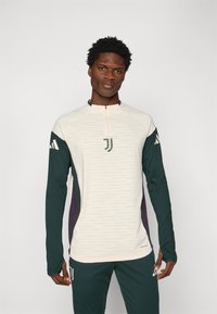 Long-sleeve athletic top in cream, green, and dark purple. Features a half-zip, textured fabric, and logo accents on the chest and sleeves.