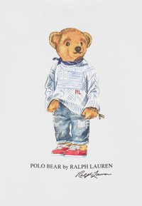 Medvědí postava v modro-bílém pruhovaném svetru s motivem americké vlajky, modrých džínech a červených teniskách, umístěná na bílém pozadí.