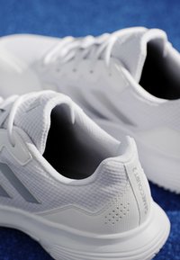 Pair of white athletic shoes with mesh texture and laces, placed on a blue surface, showing heel and side details.