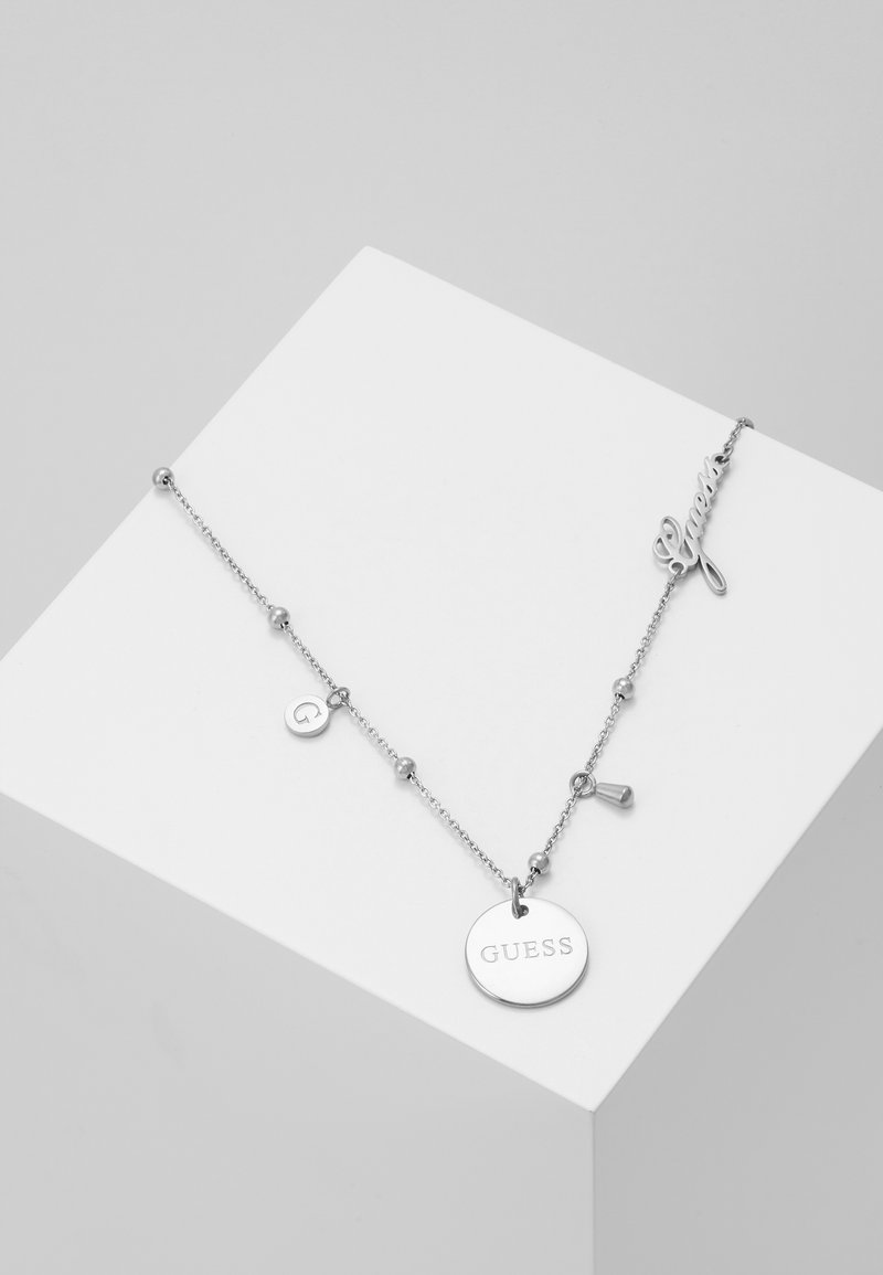 Collier chaîne ton argent avec pendentif rond "GUESS", petit charm avec "G", un charme conique et détail du script "Guess" sur une surface blanche.