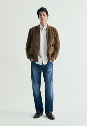 Junger Mann steht mit Händen in den Taschen, trägt eine braune Wildlederjacke, ein weißes Hemd, blaue Jeans und dunkelbraune Schuhe auf weißem Hintergrund.