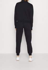 Svart fleece-träningsdräkt med hög krage, ribbade manschetter och midja; åtsittande joggingbyxor med sidofickor och ankle cuffs; i kombination med vita sneakers.