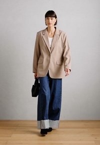 Beige Blazer mit Schulterpolstern, zwei Fronttaschen, getragen über einem weißen Hemd. Weit geschnittene Denim-Jeans mit ausgefranstem Saum und dunkler Waschung. Schwarze Handtasche.