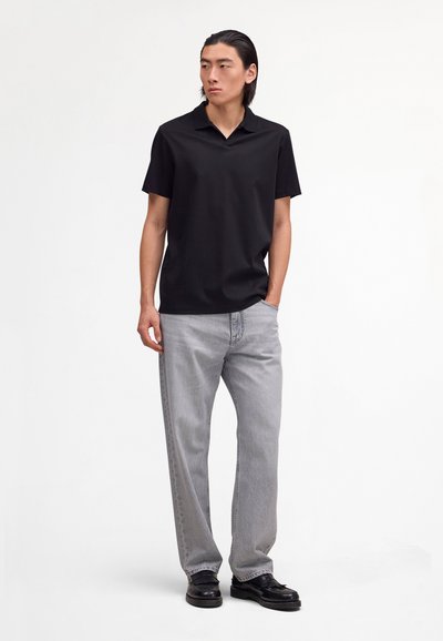 Young man standing with hands in pockets, wearing a black short-sleeve polo shirt, light gray jeans, and black shoes against a plain background.