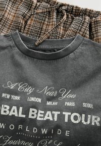 Grå T-shirt med rund halsudskæring, der har hvid tekst med "Global Beat Tour" og bynavne; parret med en brun og sort rutet nederdel.
