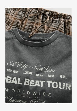 Szary t-shirt z graficznym nadrukiem i białym tekstem, na którym wymienione są miasta: Nowy Jork, Londyn, Mediolan, Paryż, Seul, zestawiony z brązowymi spodniami w kratkę na sznurki.