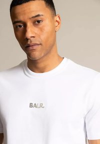 Biała koszulka z okrągłym dekoltem, wykonana z miękkiej bawełny, z srebrnym logo "BALR." umieszczonym na środku klatki piersiowej. Krótkie rękawy i gładka tekstura.