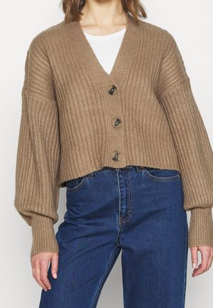 Femme portant un cardigan court côtelé camel marron avec trois boutons, des manches bouffantes et un jean bleu taille haute.
