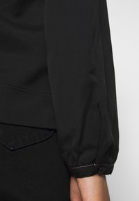 Chemise noire avec une coupe ample, des manches ballon et des poignets à boutons. Présente des coutures visibles et une texture de tissu lisse.