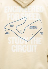Μπεζ φούτερ με κουκούλα με λευκό κείμενο "Σχεδιασμένο για ταχύτητα, μελέτησε την πίστα" και μπλε περίγραμμα χάρτη της πίστας του Αυστριακού Γκραν Πρι στο Red Bull Ring.