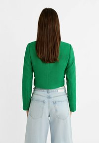 Kvinna med långt brunt hår som bär en grön kort jacka och ljusblå högmidjade jeans, stående med ryggen mot en vit bakgrund.