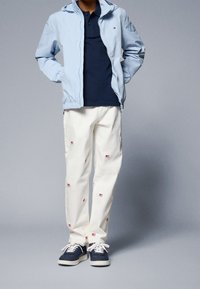 Světlá modrá lehká bunda, tmavě modré polo, bílé kalhoty s červenými vyšívanými vzory a tmavě modré tenisky s bílými tkaničkami.