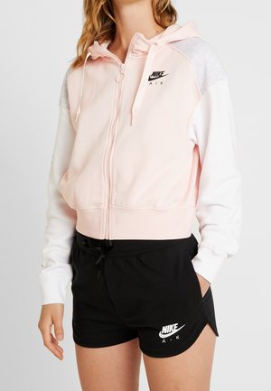 Person wearing light pink and white Nike Air zip-up hoodie and black Nike Air shorts with hands relaxed by their sides.