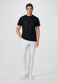 Camiseta de algodón negra con un diseño de logo gris en el centro. El modelo lleva pantalones blancos y zapatillas blancas, de pie frente a un fondo neutro.