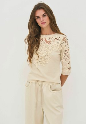 Pitkät ruskeat hiukset omaava nainen pukeutuu beigeen toppiin, jossa on kukkakuvioista pitsiä edessä ja hihoissa, yhdistettynä löysiin beigen värisiin housuihin.