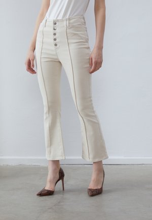 Vrouw die crèmekleurige, hooggetailleerde broek draagt met knopen aan de voorkant en bruine hoge hakken, staand op een grijze betonnen vloer.