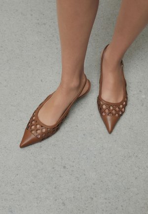 Pieds portant des ballerines slingback marron à bout pointu avec un motif découpé en treillis sur un sol texturé gris.