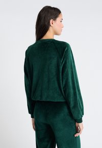 Felpa verde in velluto a coste con maniche lunghe e polsini a costine, caratterizzata da una texture morbida e una vestibilità rilassata, abbinata a pantaloni coordinati.