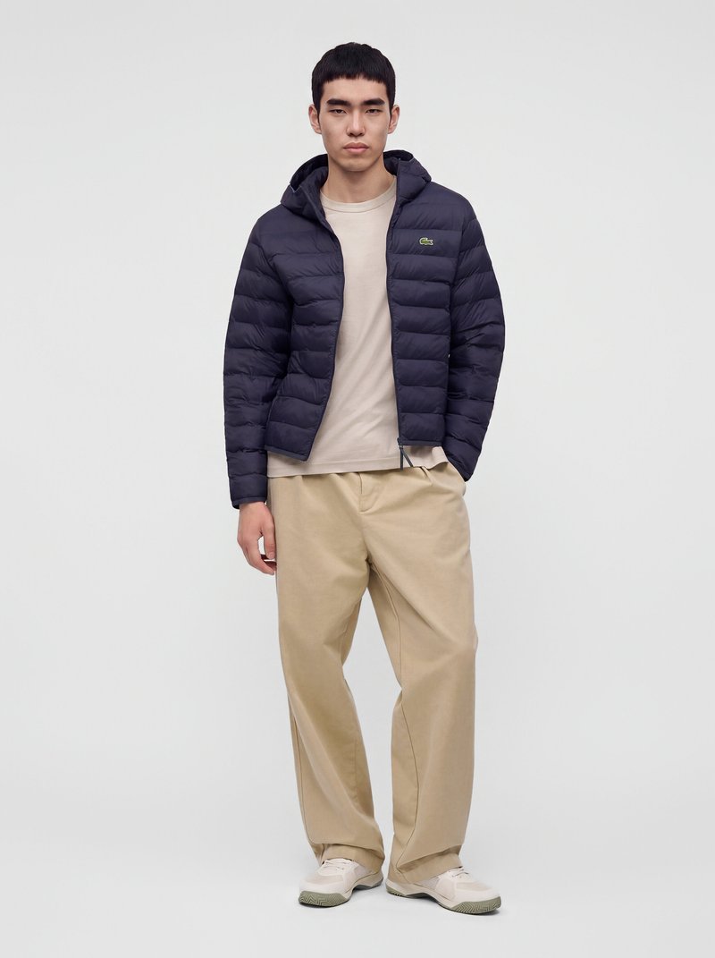 Jeune homme portant une doudoune bleu marine avec capuche, un t-shirt beige, un pantalon ample kaki et des baskets blanches, debout devant un fond uni.