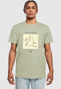 Lys grøn t-shirt med et Picasso-design af en due, med sorte konturer og typografi, der siger "PICASSO" og "FREDENS DUE 1949."