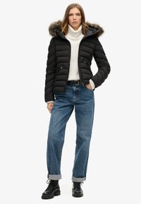 Zwarte pufferjack met een bontgevoerde capuchon, gewatteerde voorkant, geribde manchetten, gedragen over een witte gebreide trui en blauwe baggy jeans. Zwarte veterschoenen.
