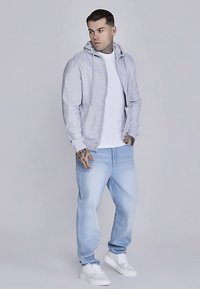 Grijze hoodie met ritssluiting, wit T-shirt eronder, lichtblauwe spijkerbroek en witte sneakers. Heeft een losse pasvorm en verstelbare capuchon.