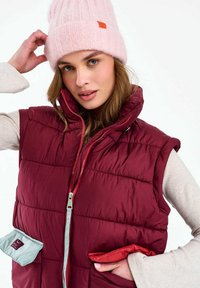 Nő pink kötött sapkát igazít, bevállig érő bordó steppelt mellényt visel bézs hosszú ujjú felső fölött, kezét a mellény zsebében tartva.