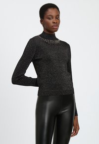 Suéter negro de cuello alto con hilos metálicos, con un escote adornado con cuentas y mangas largas. Combinado con pantalones de cuero negro.