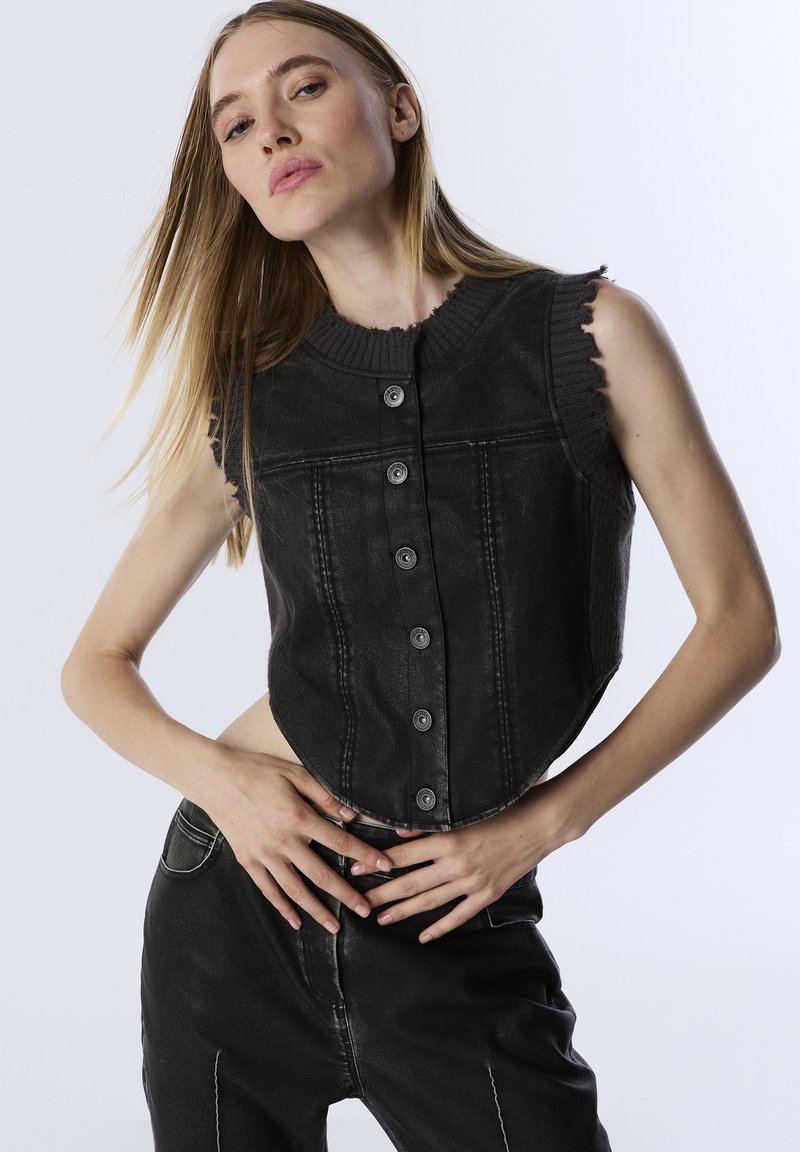 Černá zkrácená vesta vyrobená z denimu, s ribbovaným výstřihem, pěti předními knoflíkovými uzávěry a roztrhanými rukávy, doplněná o tmavé kalhoty.