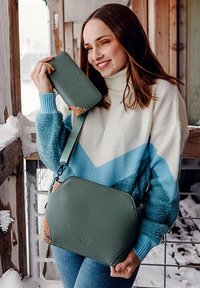 Žena v modro-bílé svetré drží zelenou kabelku a odpovídající peněženku na zasněžené dřevěné terase, usmívá se a dívá se ven.