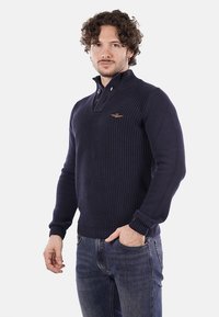 Pullover lavorato a maglia blu navy con collo a zip a un quarto, polsini a costine e un logo sul petto, abbinato a jeans blu.
