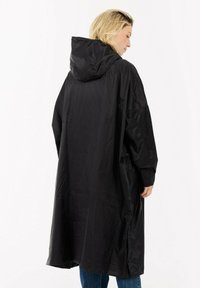 Vrouw met blond haar, gekleed in een lange zwarte regenjas met capuchon en blauwe spijkerbroek, die met haar rug naar de camera staat tegen een effen witte achtergrond.