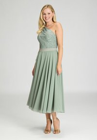 Robe vert menthe avec des détails en dentelle sur le corsage ajusté, un col asymétrique et une jupe plissée ample, complétée par des sandales à talons dorés.