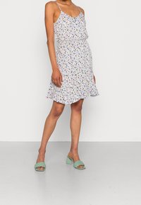 Nainen pukeutuneena hihattomaan, kukalliseen mekkoon, jossa on vyötärösolmu, ja mintunvihreisiin punottuihin korollisiin sandaaleihin, seisoo yksinkertaisella lattialla.