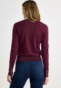 Vrouw met schouderlang haar draagt een aansluitende, mouwlange bordeauxrode trui met zilveren knopen op de manchetten en donkerblauwe spijkerbroek, gezien van achteren.