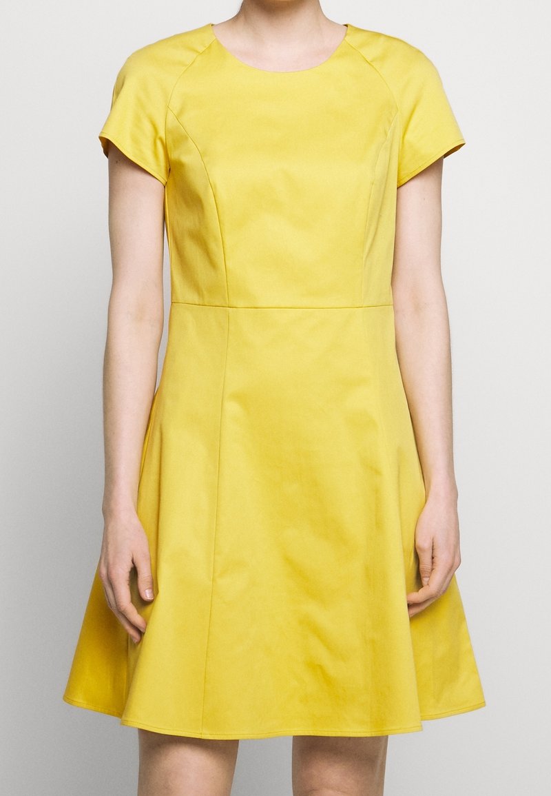 Vrouw die een gele jurk draagt met korte mouwen, tot aan de knie, met een nauwsluitend lijfje en een uitlopende rok, staand tegen een effen achtergrond.