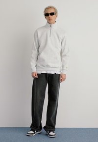 Grå lynlås sweatshirt med en frontlomme, parret med løse sorte denimbukser og sort-hvide sneakers. Modellen bærer solbriller.