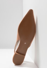 Chaussure plate beige à bout pointu reposant contre un coin blanc, montrant une semelle brune lisse avec le numéro de taille 37 en blanc.