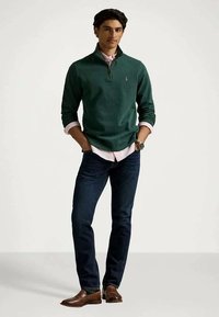 Jeune homme portant un pull vert à fermeture éclair, une chemise rose à boutons, un jean foncé, des mocassins marron, se tenant avec les mains dans les poches.